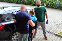 policjanci prowadzą zatrzymaną kobietę, która ma na przegubach dłoni kajdanki do samochodu służbowego