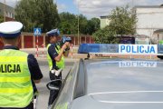 policjant ruchu drogowego stojący z drugim policjantem przy radiowozie mierzy radarem prędkość poruszających się pojazdów