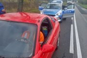 na zdjęciu widoczny na pierwszym planie czerwony samochód za kierownica którego  siedzi mężczyzna ubrany w jaskrawą bluzę z tyłu widoczny oznakowany radiowóz i policjant z niego wysiadający.