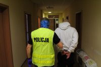 policjant w kamizelce odblaskowej prowadzi korytarzem budynku zatrzymanego mężczyznę ubranego w białą bluzę z kapturem, który na rękach trzymanych z tyłu ma nałożone kajdanki