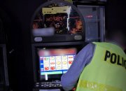 mężczyzna w odblaskowej kamizelce z napisem na plecach policja przeprowadza oględziny automatu do gier. na ekranie automatu widoczne kolorowe rysunki