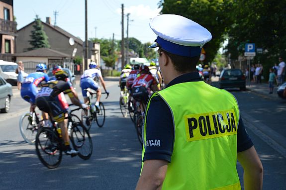 umundurowany policjant ruchu drogowego w odblaskowej kamizelce z napisem policja i czapce z białym otokiem zabezpiecza przejazd wyścigu kolarskiego. w tle widoczni kolarze jadący na rowerach