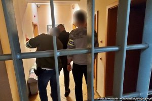 nieumundurowani policjanci / graficznie rozmyte twarze/ stoją na korytarzu za kratą policyjnego aresztu z zatrzymanym mężczyzną,  który na nadgarstki rąk trzymanych za sobą ma nałożone kajdanki