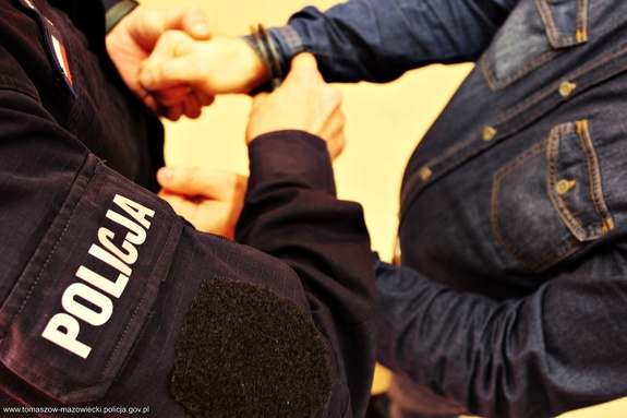 Zdjęcie poglądowe - umundurowany policjant zakłada kajdanki na przeguby dłoni mężczyzny ubranego w niebieską koszulę - sylwetki.
