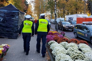 policjanci ruchu drogowego idą chodnikiem znajdującym się w rejonie cmentarza. Wokół dużo kwiatów doniczkowych na straganach