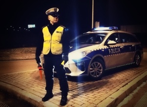 policjant ruchu drogowego w kamizelce odblaskowej i białej czapce stoi przy radiowozie