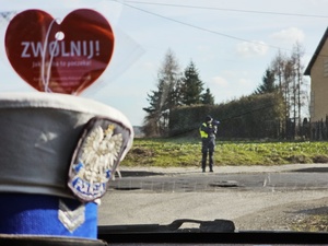 policjant ruchu drogowego mierzy prędkość nadjeżdżającego pojazdu widoczne serce z napisem zwolnij i czapka policjanta ruchu drogowego