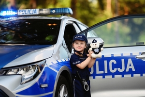 Dziecko przebrane w mundur policyjny stoi przy radiowozie z otwartymi drzwiami i w ręku trzyma policyjną maskotkę misia