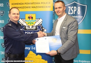Komendant Powiatowy Policji w Tomaszowie Mazowieckim inspektor Szymon Herman i Dyrektor Zespołu Szkół Ponadgimnazjalnych nr 8 w trakcie podpisywania dokumentu porozumienia. W tle baner szkoły i baner policji.