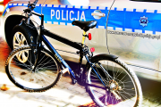 rower w czarnym kolorze a w tle oznakowany radiowóz w kolorach srebrnym i niebieskim