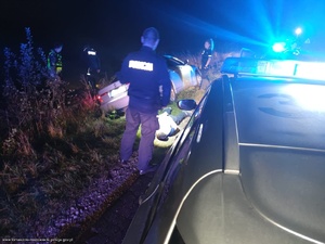 zdjęcie z miejsca zatrzymania sprawcy ucieczki przed policyjną kontrolą. Na zdjęciu widoczne dwa radiowozy  z włączoną sygnalizacją świetlną i pięciu policjantów. w części centralnej w przydrożnym rowie przechylone na lewą stronę srebrne auto osobowe marki BMW a przy jego prawym tylnym kole na ziemi leżący zatrzymany mężczyzna, oświetlony  latarką przez policjanta