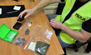 na zdjęciu widoczny policjant w kamizelce odblaskowej na blacie stołu rozkłada zabezpieczone przy przeszukaniu, torebki foliowe tak zwane dilerki z suszem roślinnym i białym proszkiem w ich wnętrzu