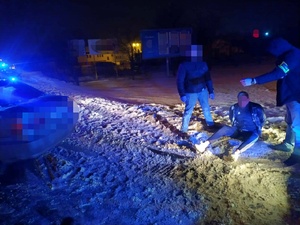 zdjęcie, po lewej stronie auto osobowe z nieczytelnymi numerami rejestracyjnymi , po stronie prawej widocznych dwóch stojących mężczyzn . mężczyzna po prawej stronie na lewym reku z opaską i napisem policja. na ziemi siedzący trzeci mężczyzna. twarze mężczyzn niewidoczne