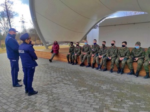 umundurowana policjantka i policjant prowadzą na wolnym powietrzu przy obiekcie amfiteatru spotkanie z grupą paramilitarną strzelec. Młode osoby biorące udział w spotkaniu są umundurowani w mundury typu wojskowego.