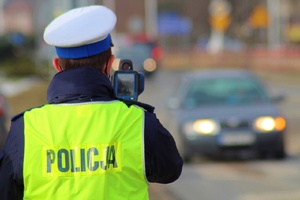 umundurowany policjant ruchu drogowego w białej czapce i odblaskowej kamizelce z napisem policja na plecach mierzy prędkość radarem pojazdu nadjeżdżającego z przeciwka. pojazd bez ostrości obrazu