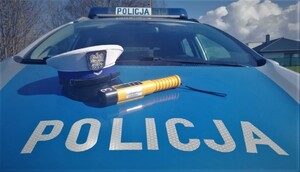 maska policyjnego radiowozu na której leży służbowa czapka z białym otokiem policjanta wydziału ruchu drogowego. obok czapki  urządzenie alcoblow do kontroli trzeźwości
