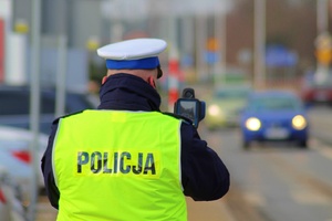 policjant ruchu drogowego w czapce z białym otokiem i odblaskowej kamizelce znapisem na plecach policja mierzy prędkość nadjeżdżającego pojazdu. Pojazd widoczny w tle - rozmyty