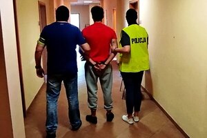 korytarz komendy, policjant i policjantka w odblaskowych kamizelkach prowadzą pomiędzy sobą zatrzymanego mężczyznę.