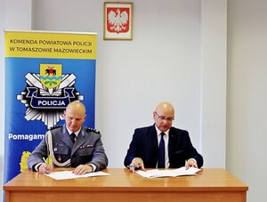 Komendant Powiatowy Policji w umundurowaniu wyjściowym wraz z Wójtem Gminy Ujazd siedząc przy biurku podpisuje porozumienie w sprawie służb ponadnormatywnych. Z tyłu widoczny policyjny baner a na ścianie godło RP