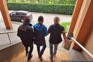 dwóch policjantów z elementami odblaskowymi z napisem policja na plecach kurtki i opasce naramiennej prowadzą między sobą zatrzymanego mężczyznę po schodach do samochodu stojącego na placu