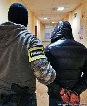 nieumundurowany policjant z opaską na parwym ramieniu z napisem POLICJA, prowadzi korytarzem zatrzymanego mężczyznę ,który na ręce trzymane za sobą ma nałożone kajdanki