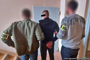 nieumundurowani policjanci / graficznie rozmyte twarze/ stoją przed drzwiami policyjnego aresztu z zatrzymanym mężczyzną,  który na nadgarstki rąk trzymanych za sobą ma nałożone kajdanki