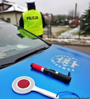 na masce radiowozu leży urządzenie do badania trzeźwości i tarcza odblaskowa tzw. lizak do zatrzymywania pojazdów. W tle widoczny policjant ruchu drogowego w odblaskowej kamizelce i białej czapce