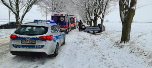 na ośnieżonej drodze stoi policyjny radiowóz a przed nim dwie karetki pogotowia. po prawej stronie od pojazdów w przydrożnym rowie leży na dachu auto osobowe a przy jego kole stoi mężczyzna