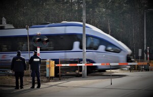 zdjęcie poglądowe przejazd kolejowy z zamkniętymi zaporami. Po torowisku porusza się pociąg. Przed przejazdem stoi umundurowany policjant i f-sz SOK