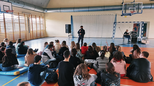 policjantka mówi do grupy kilkudziesięciu uczniów siedzących na podłodze sali gimnastycznej