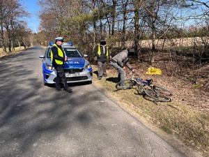 Umundurowany policjant ruchu drogowego stoi przy oznakowanym radiowozie a obok na poboczu przy lezących rowerach stoi dwóch mężczyzn, którym zasłonięto cyfrowo twarze