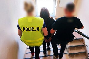 Policjantka z policjantem sprowadzają po klatce schodowej zatrzymaną kobietę idącą między nimi. Zatrzymana kobieta ma na ręce trzymane z tyłu nałożone kajdanki. Policjantka idąca po lewej stronie ma założoną odblaskową kamizelkę w jaskrawym kolorze z napisem policja