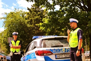 dwóch policjantów ruchu drogowego w odblaskowych kamizelkach i białych czapkach stoją przy radiowozie
