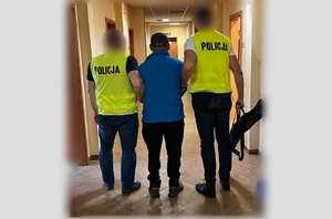 kryminalni w kamizelkach z napisem policja, prowadzą pomiędzy sobą zatrzymanego mężczyznę. policjant po prawej stronie niesie w prawym ręku pokrowiec z repliką broni odzyskaną podczas przeszukania