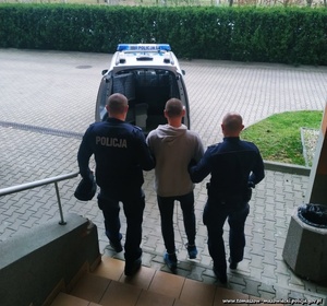 policjanci prowadzą po schodach do samochodu zatrzymanego mężczyznę, który na dłoniach i nogach ma zespolone kajdanki
