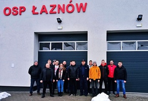 umundurowany policjant stoi przez budynkiem z napisem OSP Łaznów wraz z grupą druhów