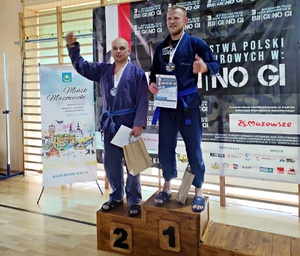 dwóch uczestników zawodów stoi na podium. Na drugim stopniu podium w niebieskim kimonie z niebieskim pasem stoi policjant z tomaszowskiej jednostki policji