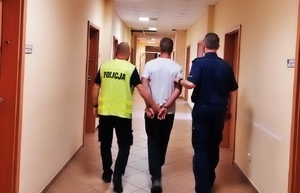 policjanci po lewej w kamizelce odblaskowej po prawo w mundurze, prowadzą pomiędzy sobą korytarzem zatrzymanego mężczyznę, który na rękach trzymanych z tyłu ma nałożone kajdanki