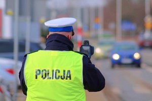 policjant ruchu drogowego w białej czapce i kamizelce odblaskowej z napisem POLICJA, mierzy prędkość laserowym miernikiem. Tło rozmyte, widoczne pojazdy