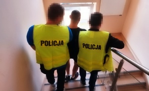 policjanci w kamizelkach odblaskowych z napisem na plecach POLICJA prowadzą między sobą, klatką schodową idąc w dół zatrzymanego mężczyznę