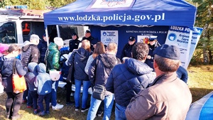 grupa ludzi stojąca przy policyjnym stoisku widoczne policyjne radiowozy, policyjny namiot i policjanci