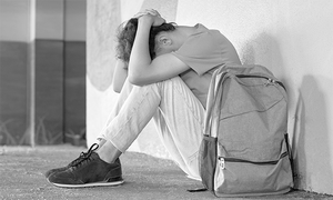 czarno białe zdjęcie, nastoletni chłopak siedzi pod ścianą z rękoma założonymi na tyle głowy po jego lewej stronie stoi plecak