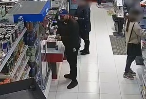 wizerunek osoby podejrzewanej o oszustwo. Mężczyzna z brodą w czarnej kurtce i spodniach oraz czapce z daszkiem na terenie sklepu