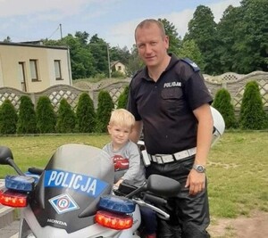 Policjant ubrany w mundur, stoi przy służbowym motocyklu, na którym siedzi mały chłopiec