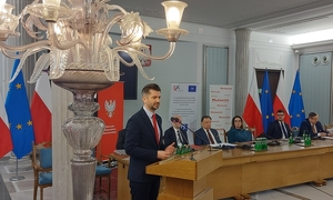 mężczyzna przy mównicy przemawia na tle uczestników konferencji. na pierwszym planie ozdobna lampa z szklanymi kloszami w oddali flagi Polski i Unii Europejskiej