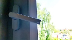 klamka okienna zostawiona w pozycji uchylnej. za szybą widoczny parking i zieleń roślinna