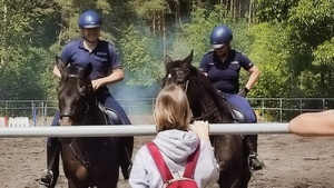 zdjęcie przedstawiające wizytę dzieci na terenie ogniwa konnego. widoczne dzieci i policyjni jeźdźcy na koniach w trakcie ćwiczeń