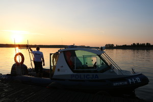 policjant stojący na pokładzie policyjnej motorówki w tle zachodzące słońce odbijające się w lustrze wody