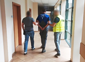 dwóch policjantów prowadzi między sobą mężczyznę z zapiętymi na ręce trzymane z tyłu kajdankami  po korytarzu komendy