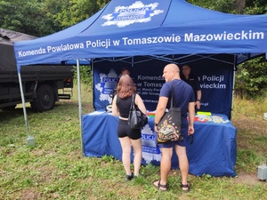 policjanci na stoisku profilaktycznym pod namiotem prezentują zainteresowanym policyjne wyposażenie służbowe oraz przekazują ulotki profilaktyczne i odblaskowe gadżety